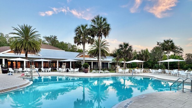 A swimming pool with lounge chairs and umbrellas, surrounded by palm trees and a building with outdoor seating. - HOT TUB & POOL TABLE The Coconut Lounge - Newly Renovated 3/2 in The Village of DeSoto (DeSoto, The Villages)