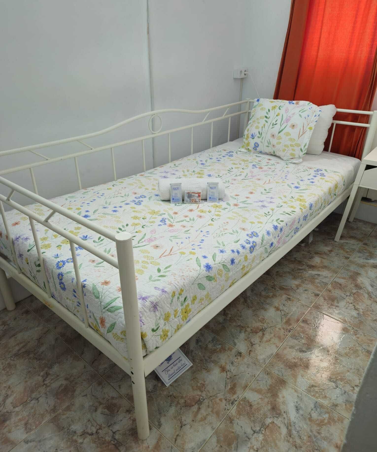 Una cama individual con colcha estampada, estructura metálica blanca y una mesita de noche con lámpara.