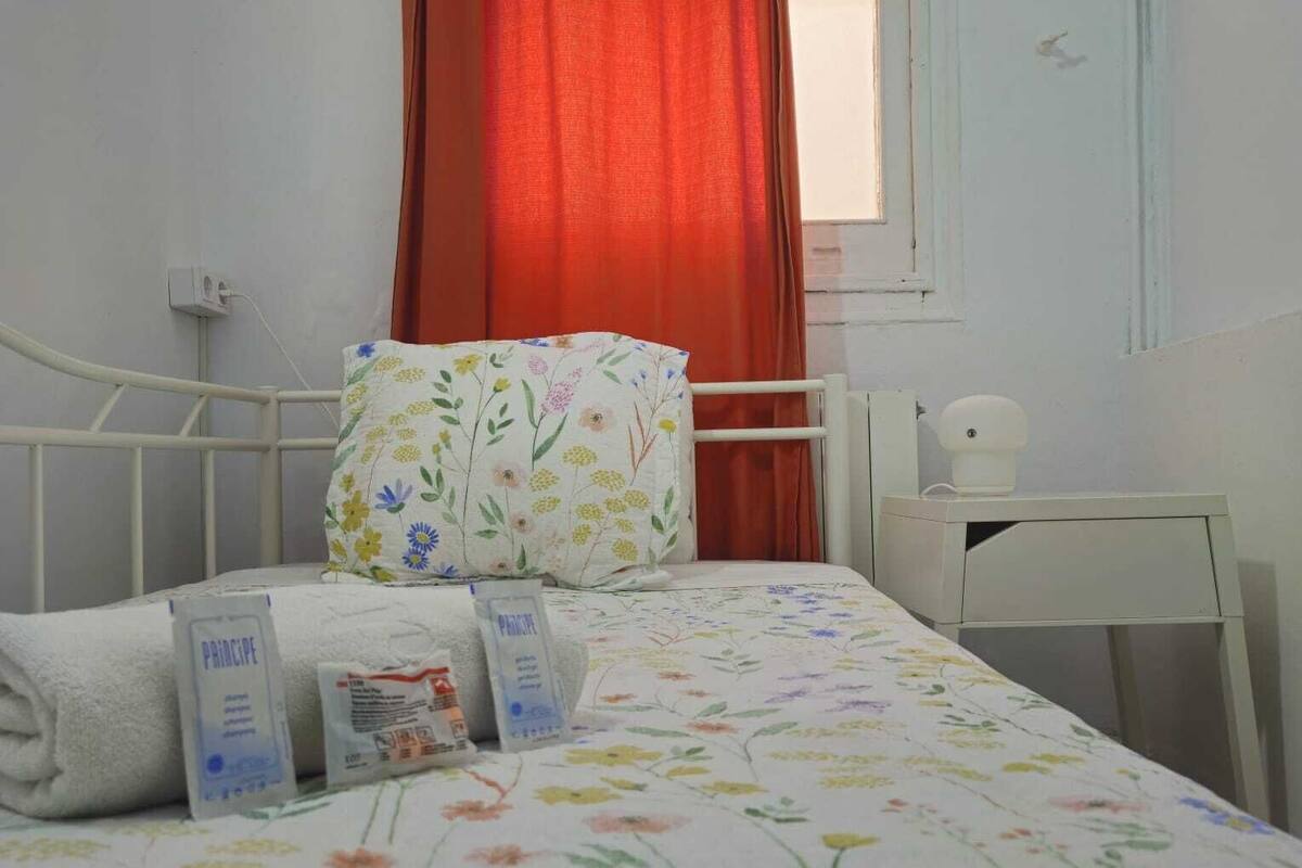 A neatly made bed with floral bedding, a white bedside table with a lamp, and a window with a red curtain.