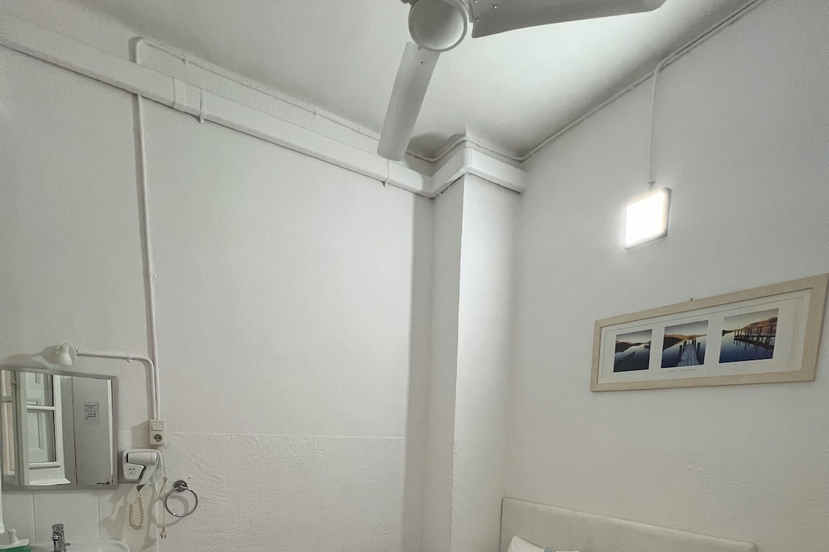 A bedroom with a bed, a ceiling fan, a bedside table, a wall-mounted light, and a framed picture.