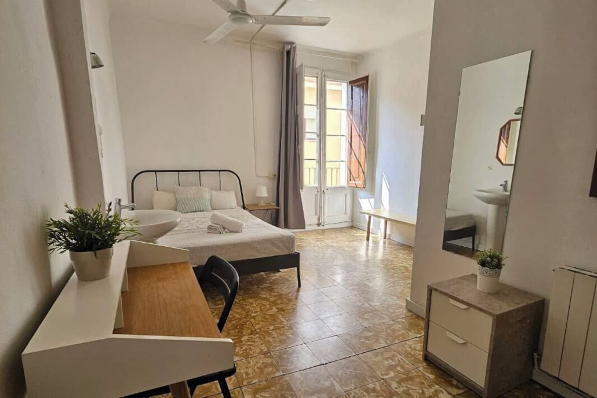 A hotel room with a bed, a desk, a chair, a small table, a mirror, and a ceiling fan.