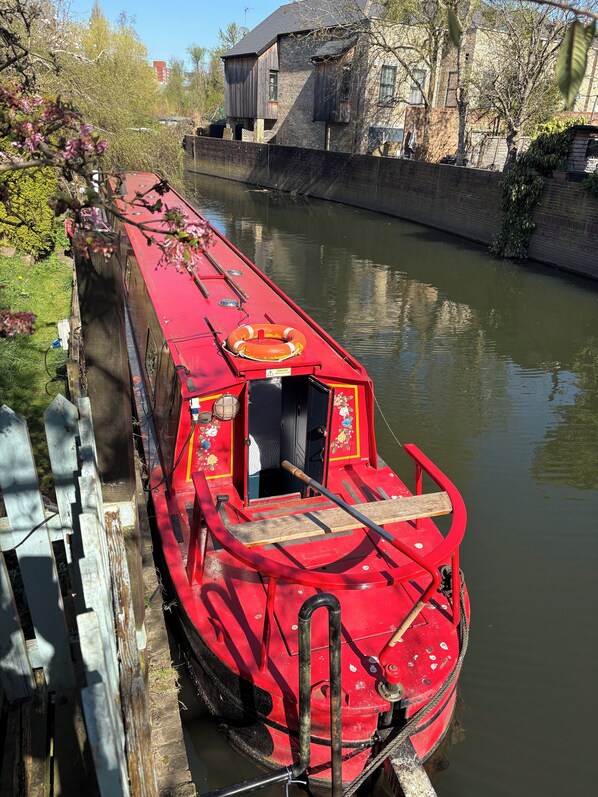 A red narrowboat docked at a canal with a wooden fence and trees in the background.