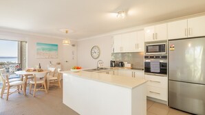 A modern kitchen with white cabinetry, a stainless steel refrigerator, a microwave, and an oven. There is a dining area with a table and chairs, and a view of the sea through a balcony.