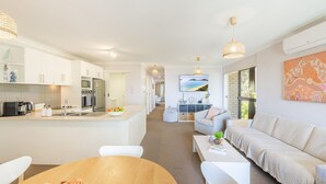 A modern kitchen with white cabinets, a built-in oven, and a coffee machine. There is a dining area with a wooden table and white chairs. The living room features a sofa and a coffee table.