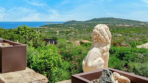Une statue de pierre dominant un paysage côtier, avec de la végétation et des collines à l’horizon.