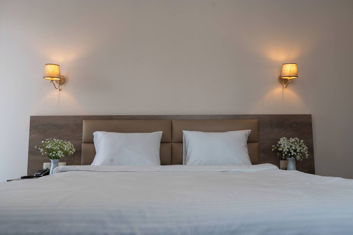 A neatly made bed with white linens, two bedside lamps, and a vase with white flowers on either side.