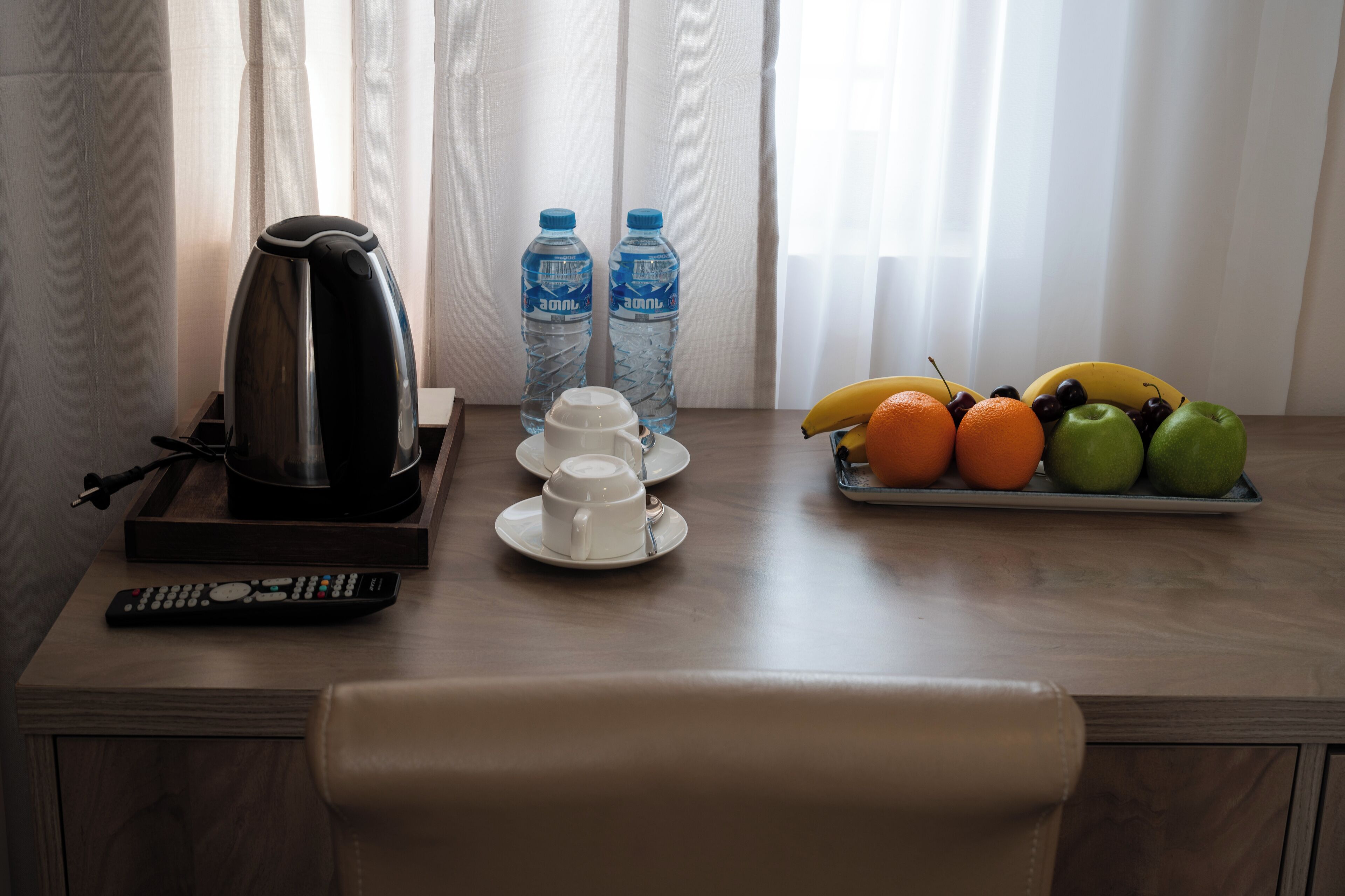 A wooden table with a kettle, two water bottles, a remote control, a cup, saucer, and a fruit tray with bananas, oranges, and apples.