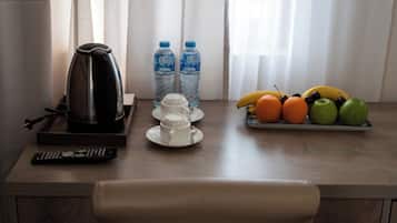 A wooden table with a kettle, two water bottles, a remote control, a cup, saucer, and a fruit tray with bananas, oranges, and apples.