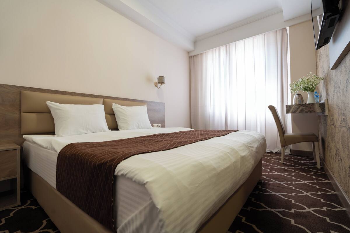A hotel room with a large bed, a nightstand, a chair, a TV, and a window with curtains.