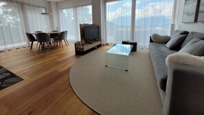 Un salón moderno con suelo de madera, un sofá gris, una mesa de centro de cristal, un televisor sobre un mueble bajo y una zona de comedor con mesa y sillas.
