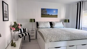 Une chambre à coucher avec un lit, deux tables de chevet, une chaise, une lampe, un tableau au mur et une fenêtre avec des rideaux.