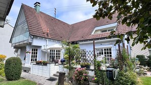 Une maison à deux étages, dotée d’un toit en pente, d’un balcon et d’un jardin agrémenté de plantes en pot et de fleurs.