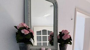 Une coiffeuse bleu clair, dotée d’un grand miroir, agrémentée de deux plantes en pot aux fleurs roses et de trois petits récipients placés sur la cheminée.