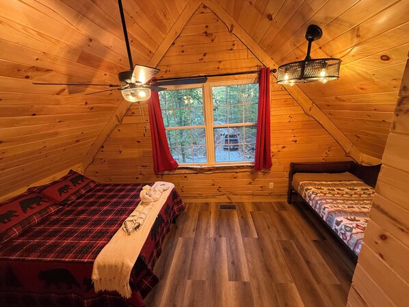 A cozy cabin room with two beds, a large window with red curtains, wooden walls, and a sloped ceiling.