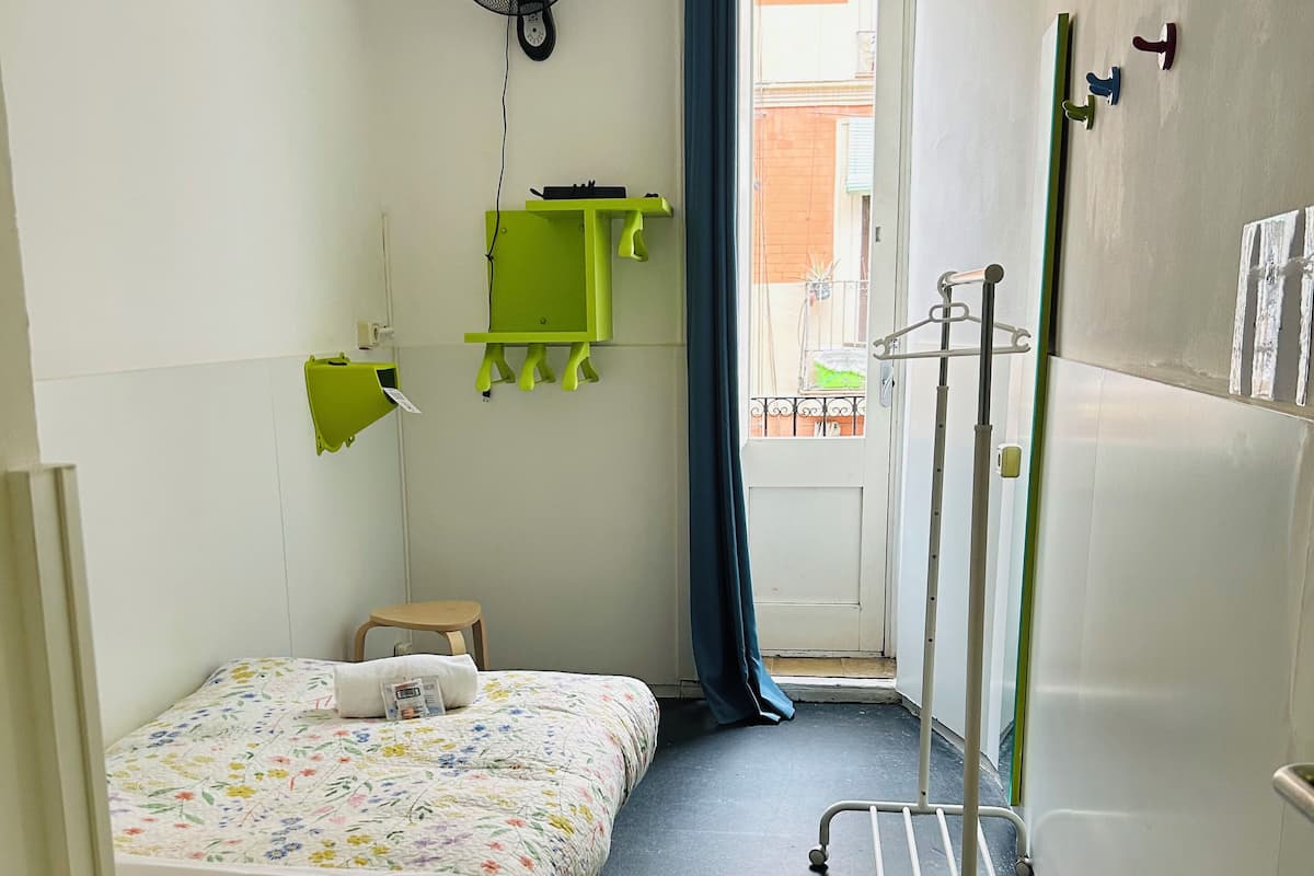 A small, narrow hotel room with a bed, a sink, a fan, and a window with a view of the outside.