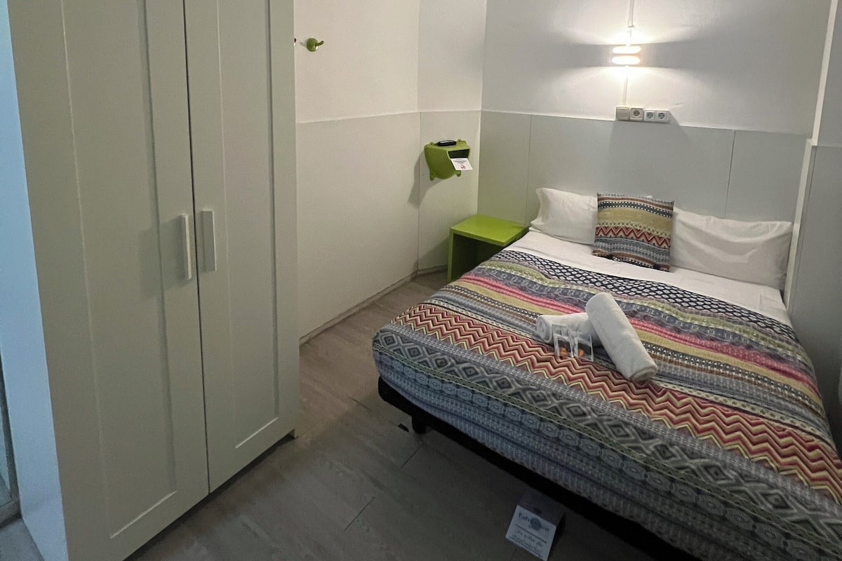 A small, well-lit bedroom with a bed, a wardrobe, a bedside table, and an air conditioning unit.