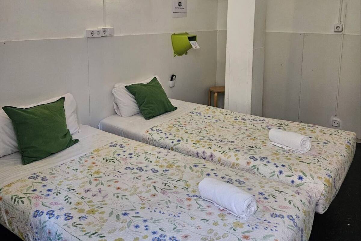 A hotel room with two beds, each with a floral-patterned bedspread and green pillows. There is a small table and a green wall-mounted phone.