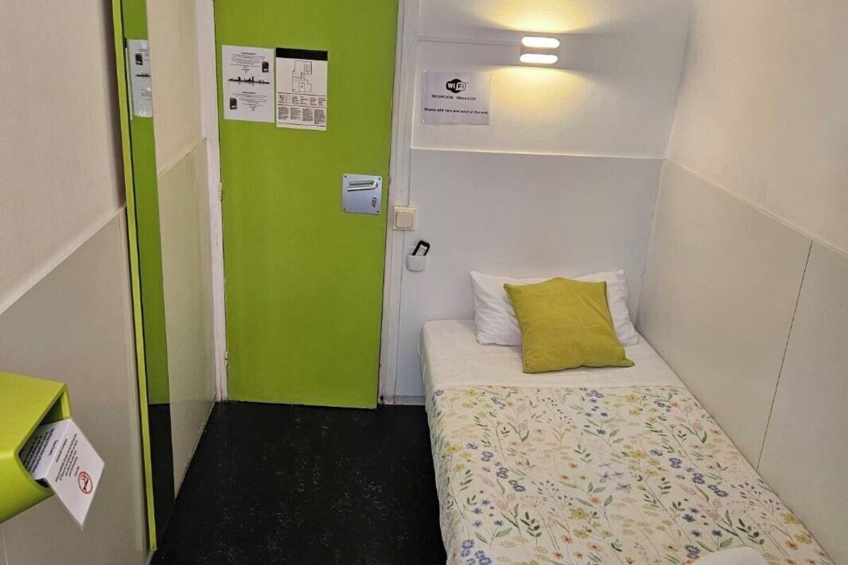 A small, narrow hotel room with a green door, a bed with a floral bedspread, a white pillow, a yellow pillow, and a wall-mounted light fixture.