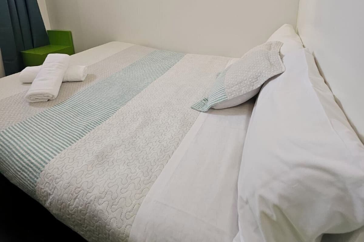 A hotel room with a neatly made bed, a green chair, and a wall-mounted light.