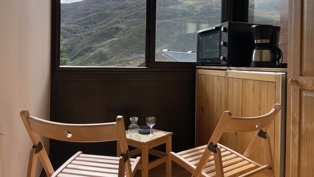 Une pièce avec vue sur un paysage montagneux, grâce à de grandes fenêtres. On y trouve une table en bois avec deux chaises, une petite table avec un verre et une tasse, et un four à micro-ondes posé sur un comptoir en bois.