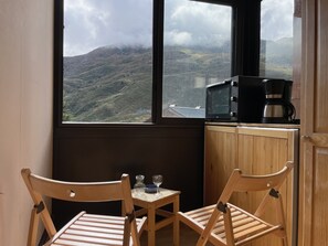 A room with a view of a mountainous landscape through large windows. There is a wooden table with two chairs, a small table with a glass and a cup, and a microwave on a wooden counter.