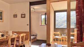 Une pièce chaleureuse avec une table à manger en bois, des chaises, un lit avec une literie à motifs, une petite table avec des tasses et un bol, et une fenêtre donnant sur la montagne.