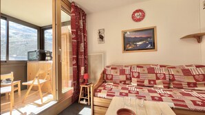 Une pièce chaleureuse avec un canapé en bois, une petite table sur laquelle se trouvent un bol et des verres, une chaise, un meuble en bois, un rideau rouge et une horloge au mur.