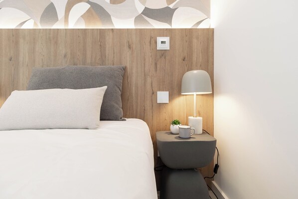 A neatly made bed with two pillows, a bedside table with a lamp, a small plant, and a power outlet.