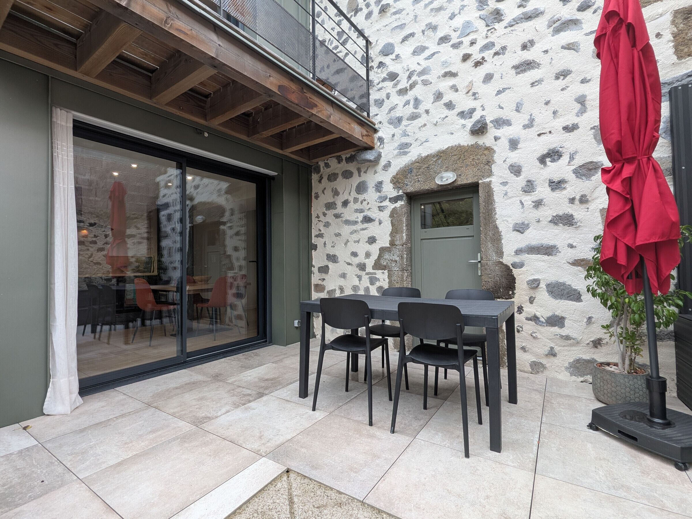 A stone exterior wall, a sliding glass door, a table with chairs, and a red umbrella.
