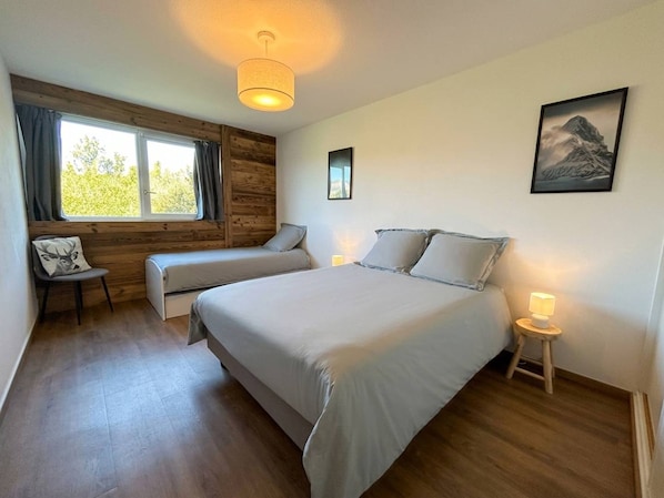 A bedroom with a large bed, a chair with a cushion, a bedside table with a lamp, and a window with curtains.