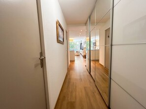 A hallway with a wooden floor leading to a room with a bed and a window.