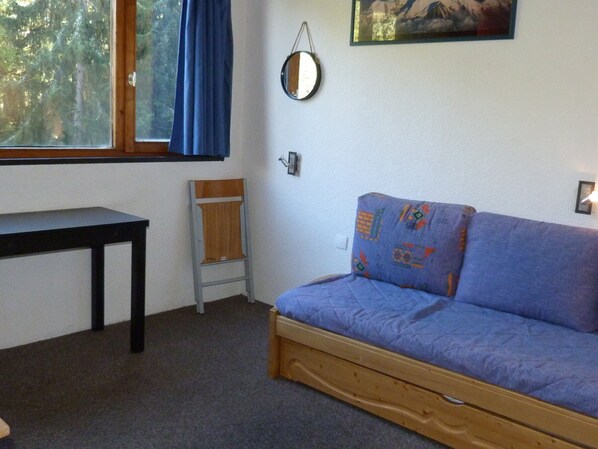 A room with a wooden bench, a blue sofa, a small table, a window with blue curtains, a wall-mounted mirror, and a painting. - Cozy studio in Arc 1800 for 2 people (Les Arcs 1800)