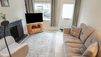 Uma sala de estar com lareira, televisão sobre um suporte de madeira, sofá bege e um tapete estampado.