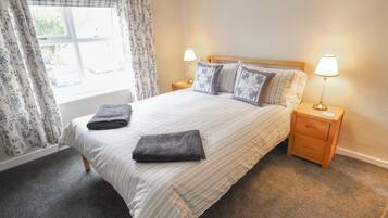 Cama bem arrumada com duas almofadas, duas luminárias de cabeceira e uma janela com cortinas.