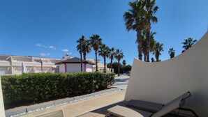 A sunlit outdoor seating area with a table and chairs, surrounded by a hedge and palm trees.
