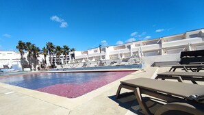 A pool area with lounge chairs and umbrellas, palm trees, and a modern building in the background.