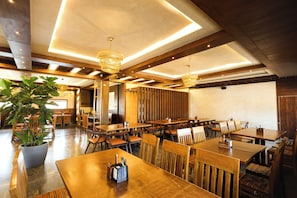 Een ruime restaurantruimte met houten tafels en stoelen, hanglampen en een grote potplant.