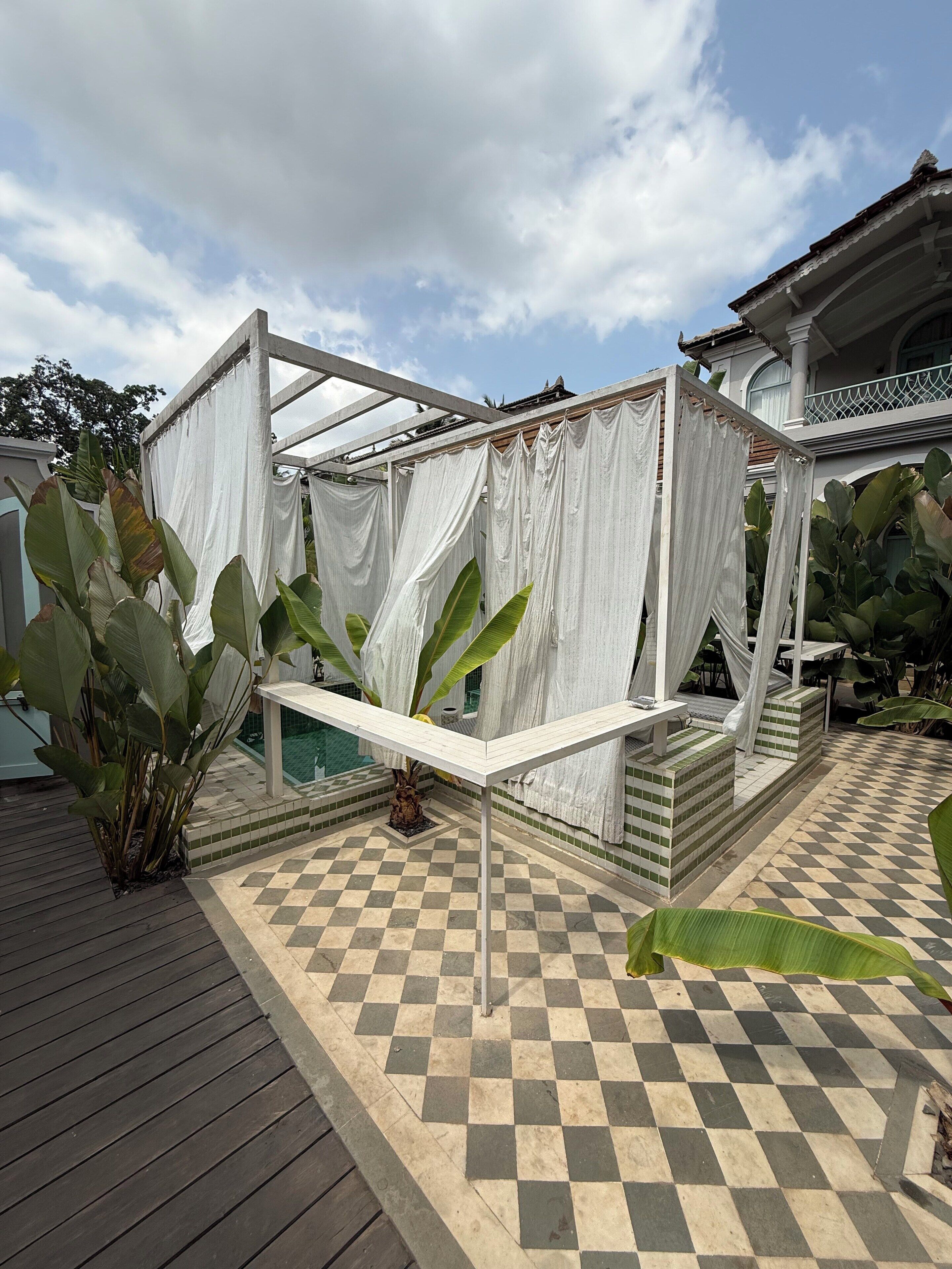 A covered outdoor seating area with a checkered floor, surrounded by large plants and a white structure.