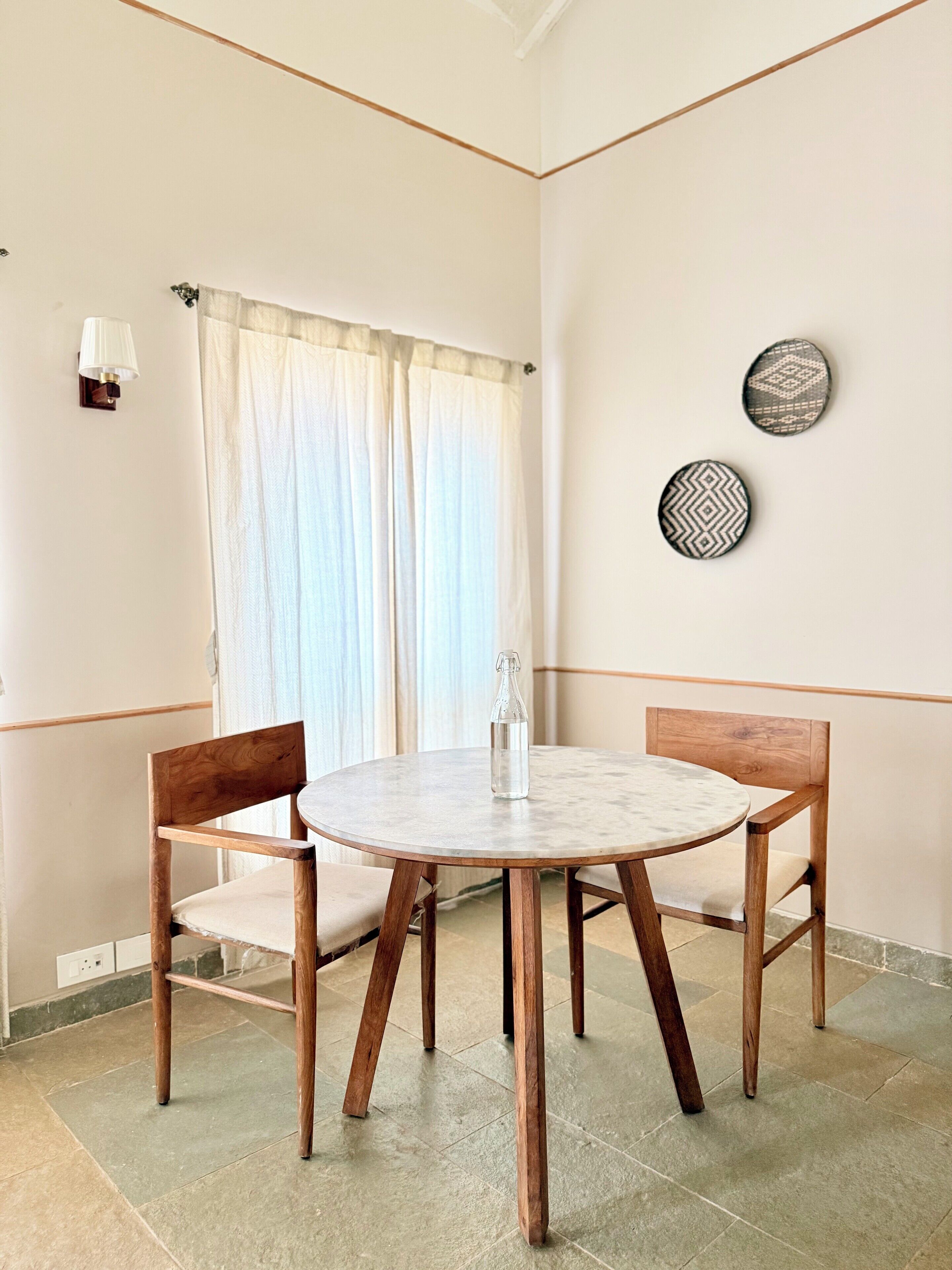 Un comedor con una mesa redonda, dos sillas de madera, una botella de agua, una ventana con cortinas y dos placas decorativas colgadas en la pared.