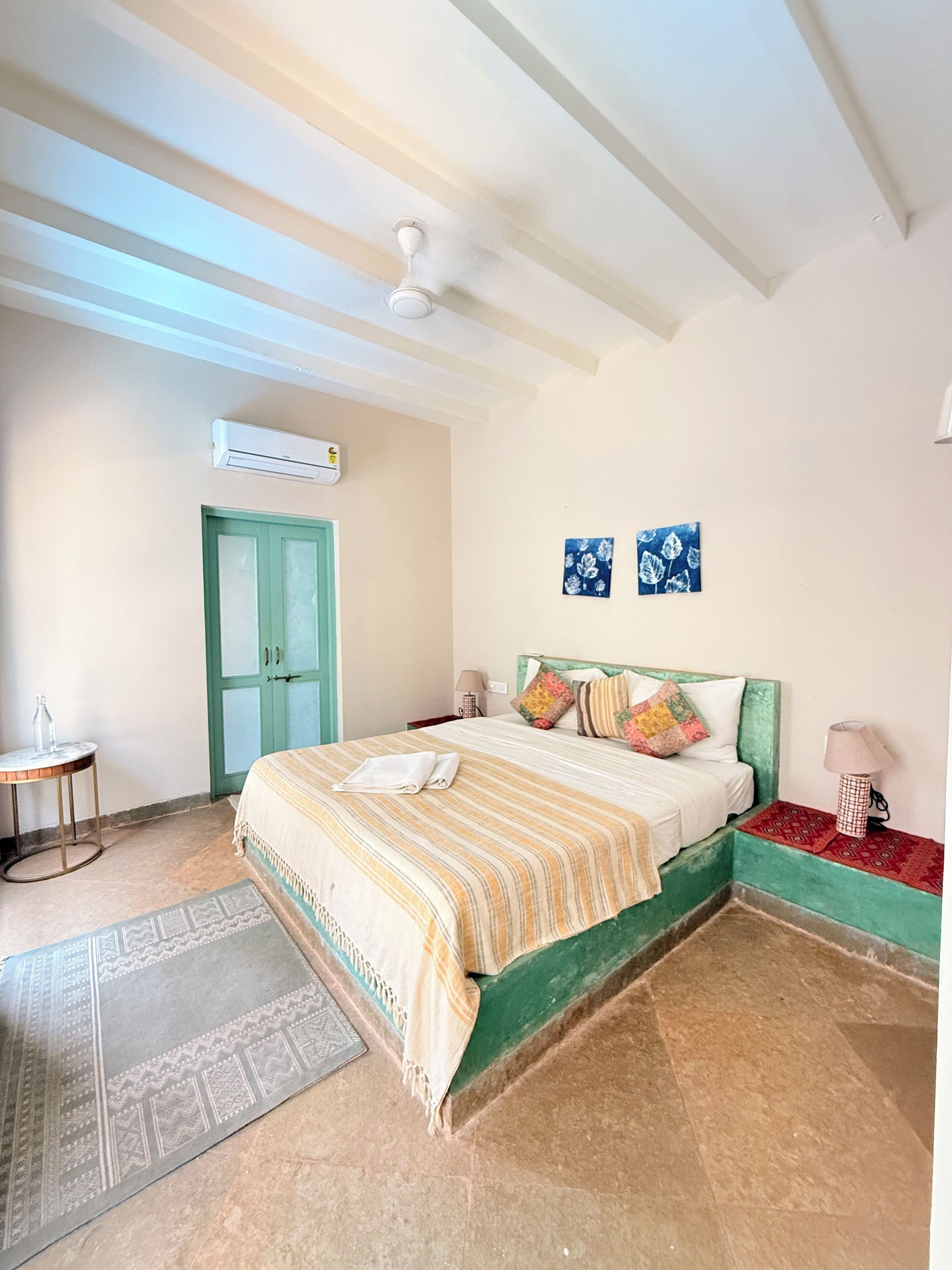 A bedroom with a bed, a green door, an air conditioning unit, a small table with a bottle, and two wall-mounted artworks.