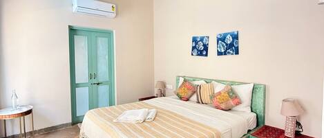 A bedroom with a bed, a green door, an air conditioning unit, a small table with a bottle, and two wall-mounted artworks.