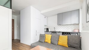 A compact living space with a kitchenette, a sofa with yellow cushions, and a wooden table. - RentPlanet - Fort Wola (Warsaw)
