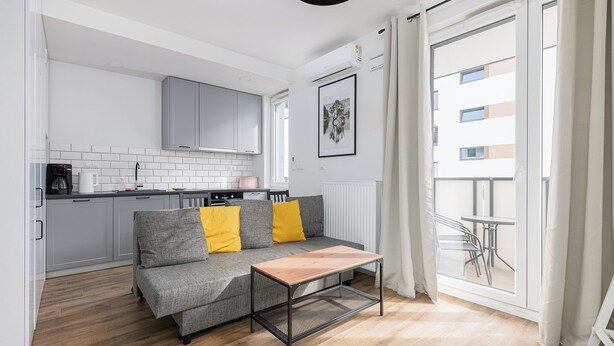 A modern living room with a grey sofa, wooden coffee table, and a view of a balcony with a table and chairs. - RentPlanet - Fort Wola (Warsaw)