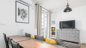 A modern living room with a dining area, a grey sofa with yellow cushions, a wooden dining table, and a grey cabinet with a flat-screen TV. - RentPlanet - Fort Wola (Warsaw)