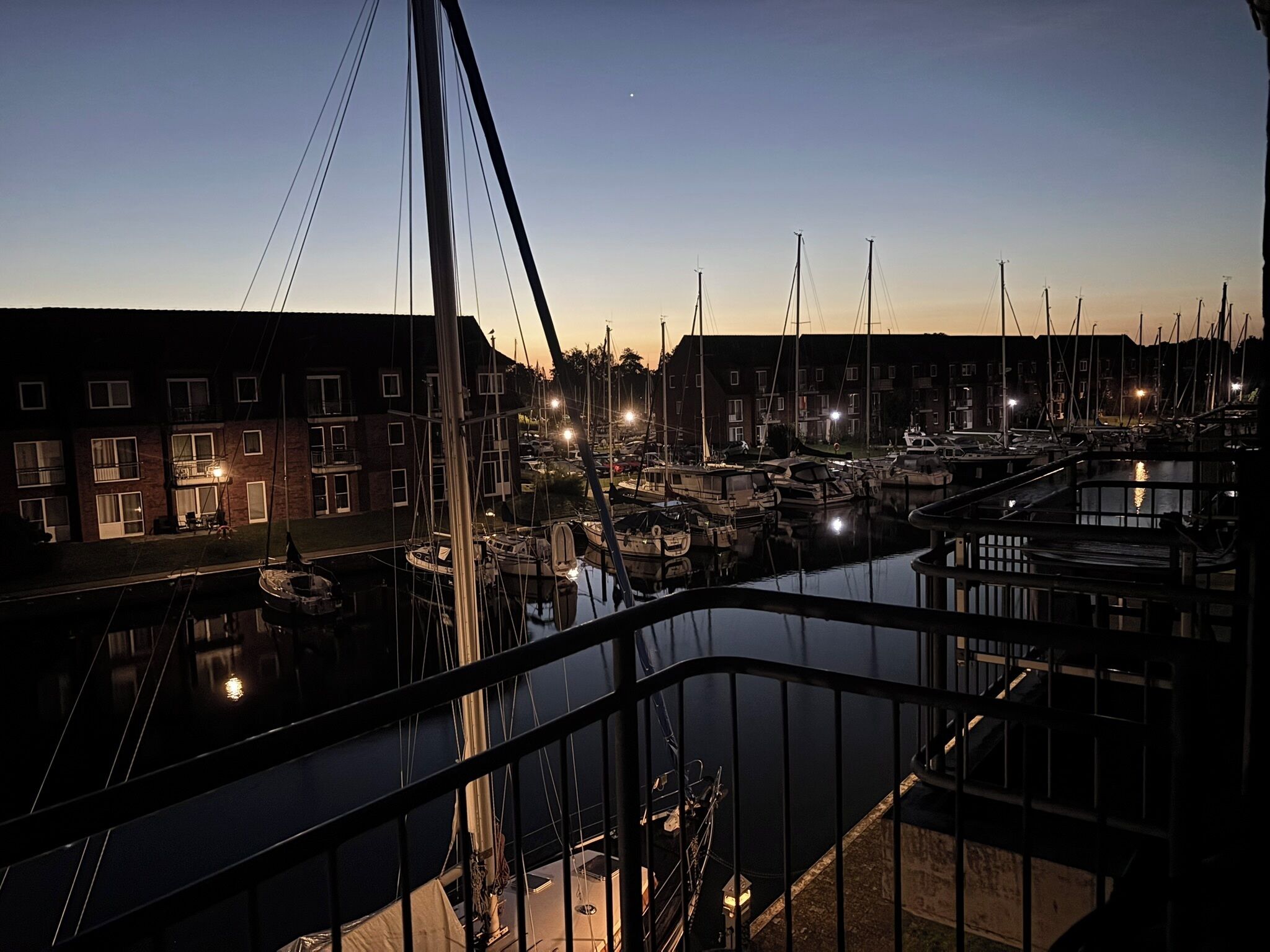 A marina at dusk with boats docked, a bridge in the foreground, and buildings in the background.