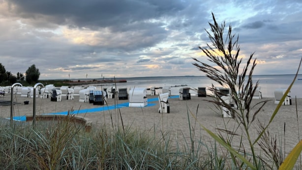 Zona de platja amb gandules i ombrel·les blanques, un camí de sorra i vista al mar sota un cel amb núvols.