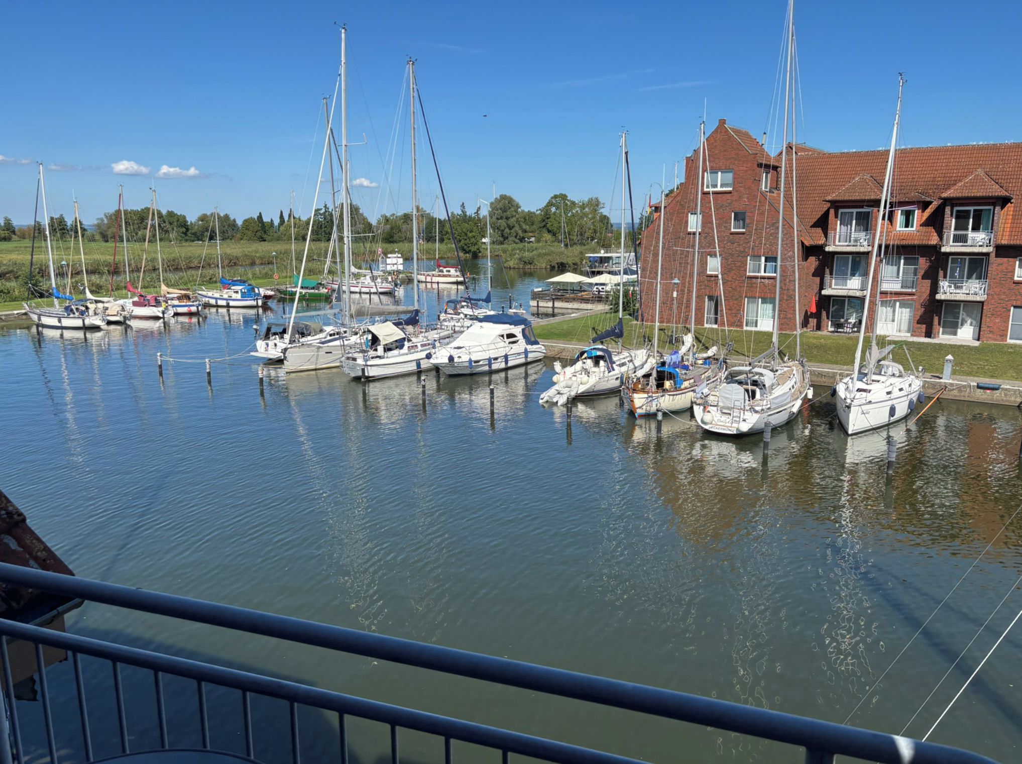 En lystbådehavn med flere sejlbåde i havnen, en murstensbygning med balkoner, og en klar blå himmel.