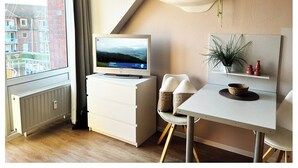 A modern room with a TV on a dresser, a dining table with chairs, a plant on a shelf, and a view of buildings through a window. - Maritime apartment in the marina of the lagoon town of Ueckermünde (Ueckermünde)