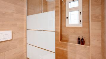Une salle de bain moderne avec un plancher en bois, une douche à l’italienne blanche, une fenêtre intégrée et deux bouteilles de shampoing posées sur une étagère.