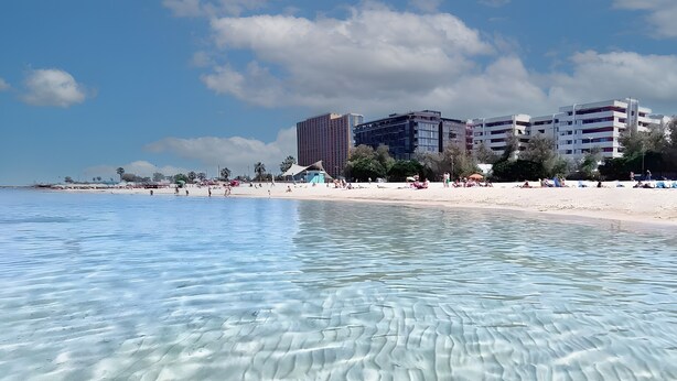 Une plage avec une eau claire, un rivage sablonneux et des bâtiments en arrière-plan.